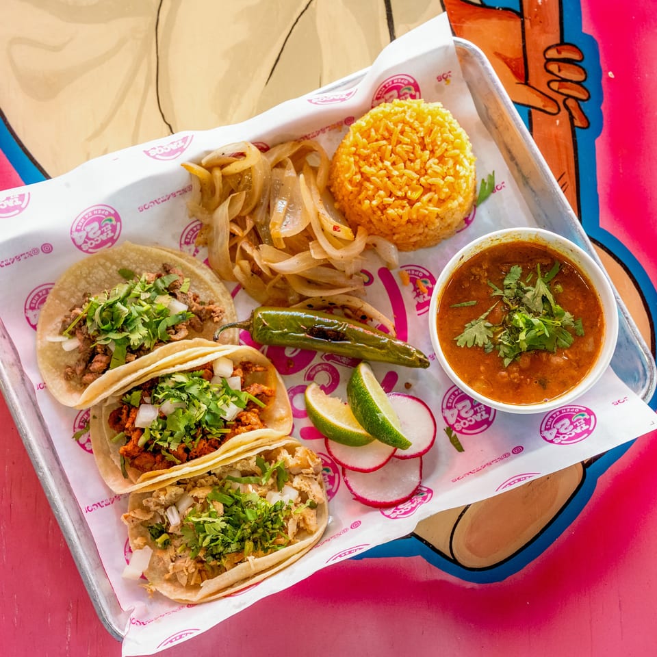 Three tacos, rice, grilled onions, a chile, limes, radishes, and a bowl of soup on a tray.