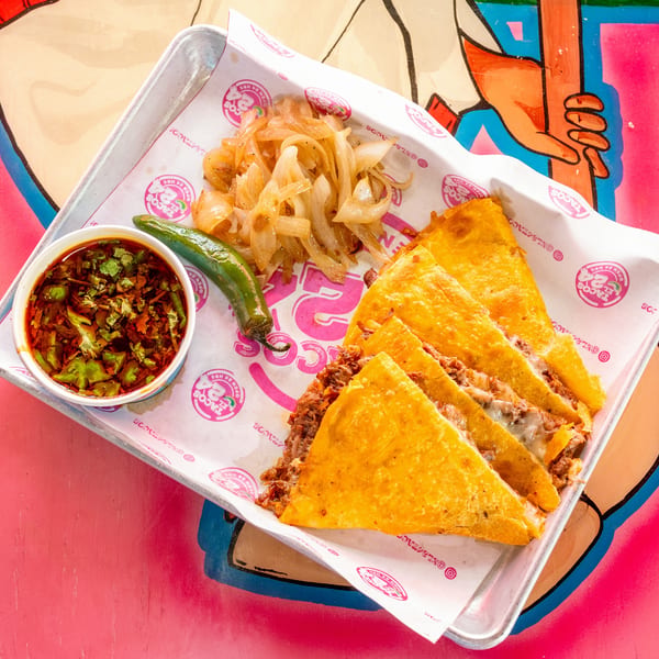 Birria quesadillas with consome, grilled onions, and a serrano pepper on a metal tray.